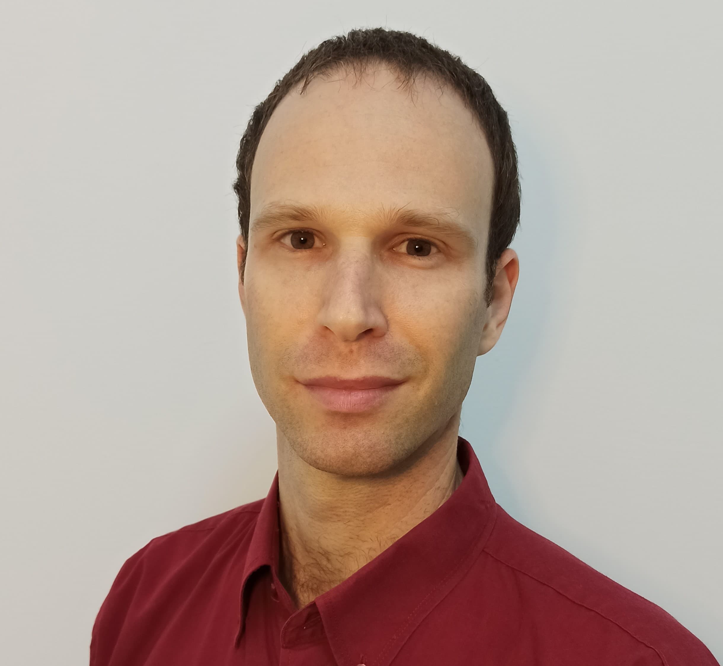 Man with short brown hair and brown eyes wearing a maroon shirt against white background.