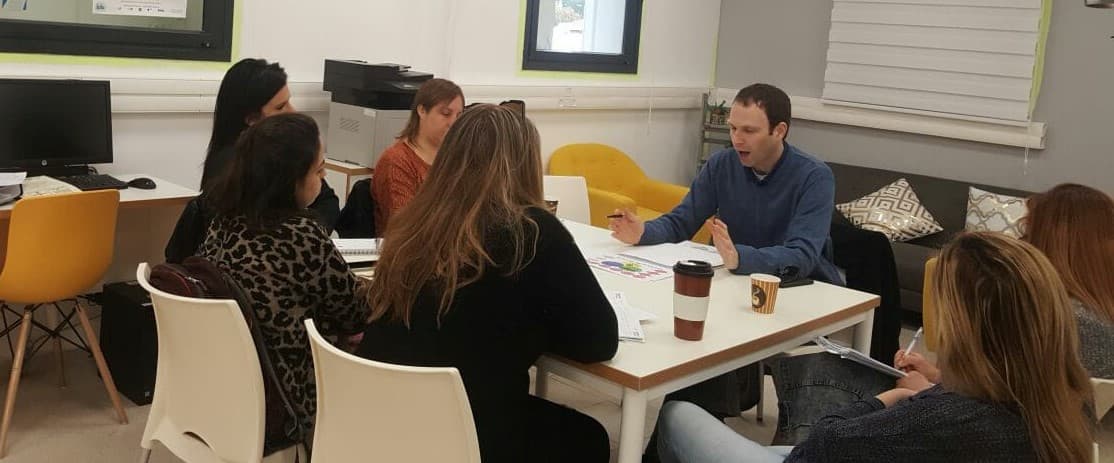 A man gestures while leading a discussion with colleagues seated around a white table.