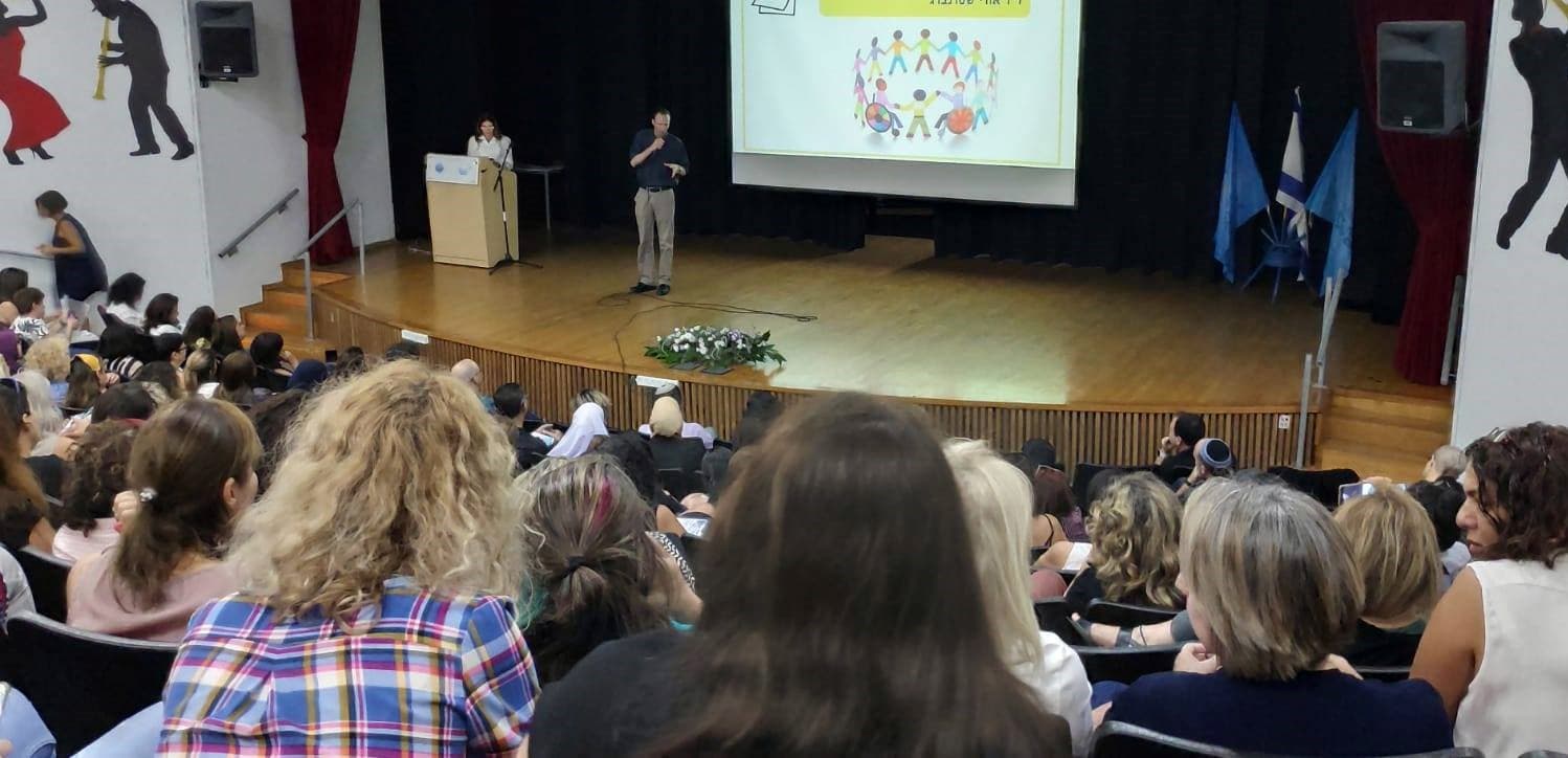 Speaker on stage addressing a seated audience in an auditorium with a large projection screen.