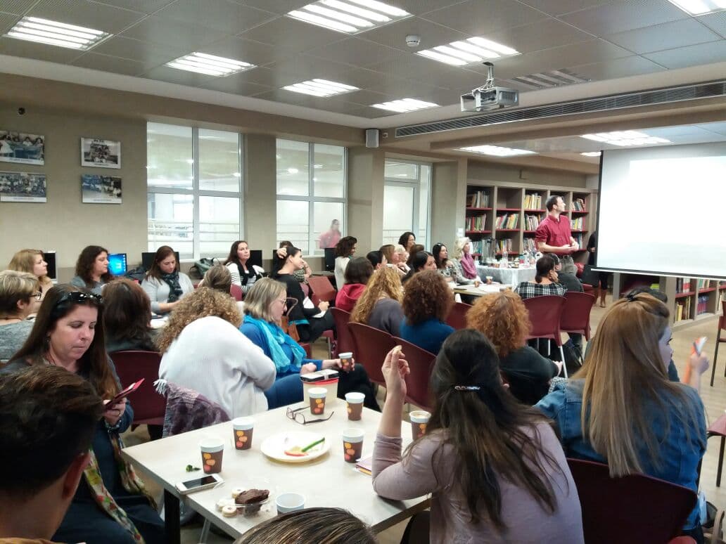 People seated at tables listen to a man presenting in a library with bookshelves.